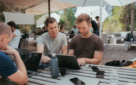 Zwei Personen unter einem grossen weissen Sonnenschrim schauen auf ein Macbook. Gegenüber sitzt eine weitere Person, die am Laptop arbeitet. Ein Smartphone liegt auf dem Tisch.