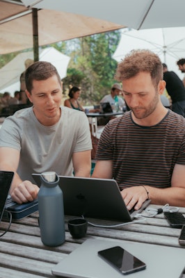 Zwei Personen unter einem grossen weissen Sonnenschrim schauen auf ein Macbook. Gegenüber sitzt eine weitere Person, die am Laptop arbeitet. Ein Smartphone liegt auf dem Tisch.