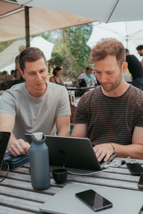 Zwei Personen unter einem grossen weissen Sonnenschrim schauen auf ein Macbook. Gegenüber sitzt eine weitere Person, die am Laptop arbeitet. Ein Smartphone liegt auf dem Tisch.