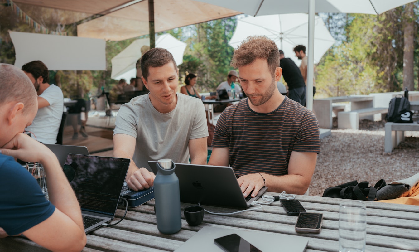 Zwei Personen unter einem grossen weissen Sonnenschrim schauen auf ein Macbook. Gegenüber sitzt eine weitere Person, die am Laptop arbeitet. Ein Smartphone liegt auf dem Tisch.