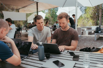 Zwei Personen unter einem grossen weissen Sonnenschrim schauen auf ein Macbook. Gegenüber sitzt eine weitere Person, die am Laptop arbeitet. Ein Smartphone liegt auf dem Tisch.