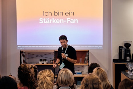 Ein Mann spricht vor einer Präsentationsfolie mit dem deutschen Text „Ich bin ein Starken-Fan“ zu einem sitzenden Publikum.