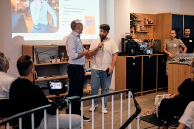 Zwei Männer stehen während einer Präsentation in einem modernen Raum mit digitalem Bildschirm und davor sitzendem Publikum und sprechen in Mikrofone. (Hinweis: Im bereitgestellten Text waren keine „ß“-Zeichen zum Ersetzen vorhanden.).