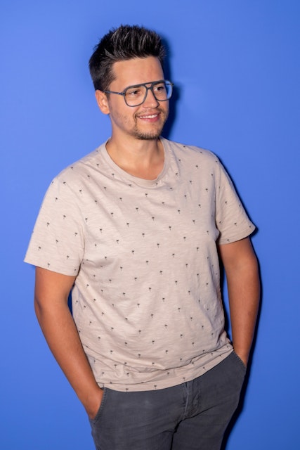 Ein Mann mit Brille, hellem gemustertem T-Shirt und dunkler Hose steht mit den Händen in den Taschen vor einem einfarbig blauen Hintergrund und lächelt leicht.