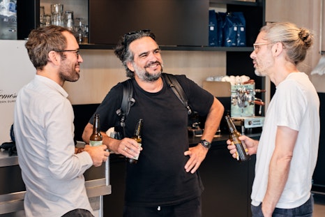 Drei Männer stehen in einer modernen Küche, halten Bierflaschen in der Hand und plaudern neben einer Kaffeemaschine miteinander.