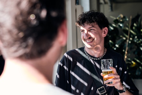 Eine Person mit einem Glas Bier in der Hand lächelt und plaudert mit einer anderen Person im Haus, während das Sonnenlicht Schatten auf ihr Gesicht wirft.