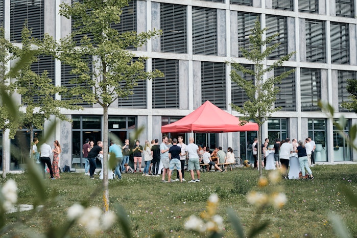 Die Menschen versammeln sich unter und um ein rotes Zeltdach vor einem modernen Gebäude mit grossen Fenstern auf einem grasbewachsenen Gelände mit jungen Bäumen.