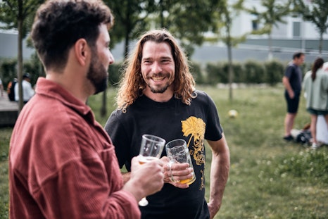 Zwei Männer stehen draussen mit Biergläsern in der Hand und unterhalten sich; der eine lächelt den anderen an. Im Hintergrund erscheinen Menschen und Bäume.