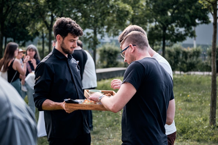 Ein schwarz gekleideter Kellner trägt ein hölzernes Tablett mit Essen, während die Leute bei einer Veranstaltung im Freien mit Bäumen im Rücken Snacks auswählen.