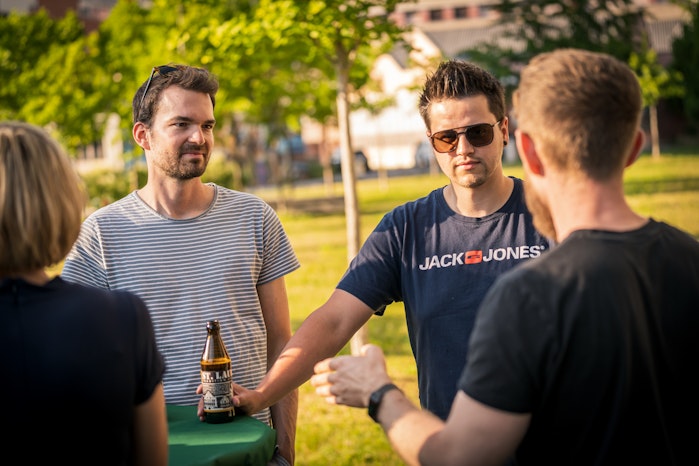 Vier Personen unterhalten sich draussen an einem grünen Tisch. Einer hält eine Bierflasche in der Hand. Es ist sonnig, mit Bäumen und Gebäuden hinter ihnen.