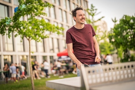 Ein Mann in einem kastanienbraunen Hemd lächelt, während er draussen in der Nähe eines modernen Gebäudes Tischtennis spielt, Menschen und Bäume hinter ihm.