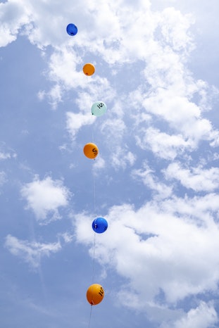 Bunte Luftballons mit Zahlen schweben in einer vertikalen Linie vor einem teilweise bewölkten, strahlend blaün Himmel nach oben.