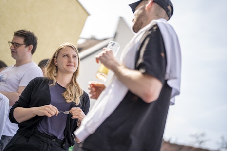 Eine Frau und zwei Männer stehen im Freien und unterhalten sich tagsüber. Ein Mann hält ein Getränk in der Hand, das Hemd über der Schulter. Gebäude im Hintergrund.