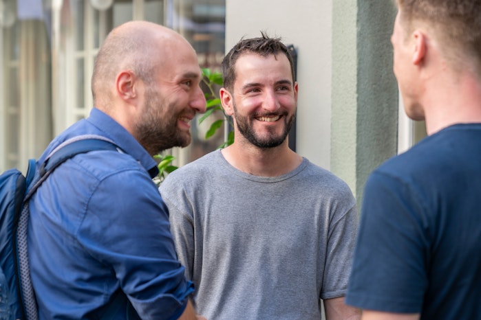 Drei Männer stehen draussen, lächeln und unterhalten sich. Einer trägt ein blaüs Hemd mit Rucksack, einer ein graüs T-Shirt und einer ein dunkles Hemd von hinten gesehen.