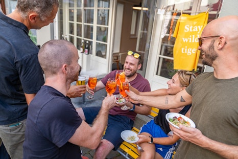 Fünf Menschen stossen im Freien mit Gläsern an, lächeln und unterhalten sich bei Essen und Getränken auf einer gelben Bank in der Nähe eines Gebäudes.