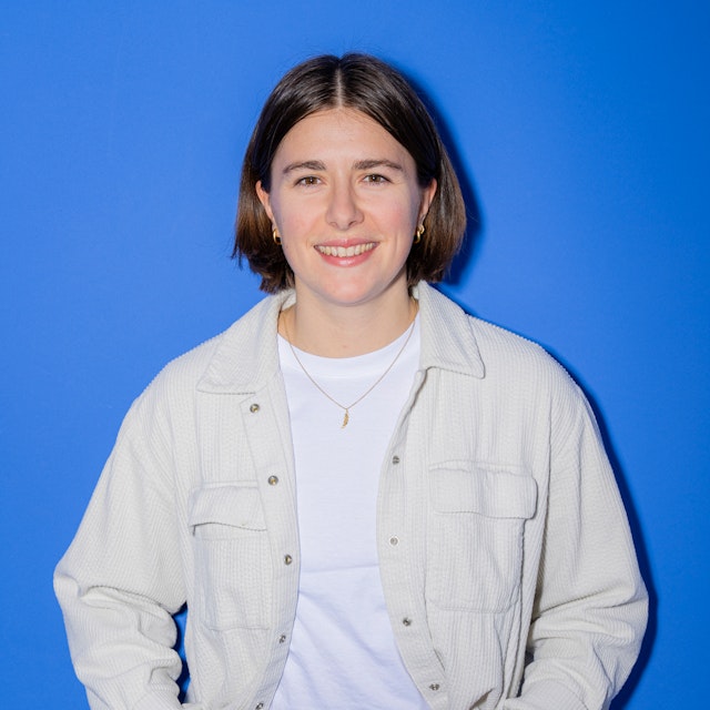 Eine Person mit kurzen braunen Haaren, einem weissen Hemd, einer hellen Jacke, einer schwarzen Hose und einer Uhr steht lächelnd vor einem blaün Hintergrund.