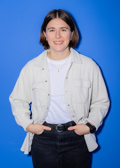 Eine Person mit kurzen braunen Haaren, einem weissen Hemd, einer hellen Jacke, einer schwarzen Hose und einer Uhr steht lächelnd vor einem blaün Hintergrund.