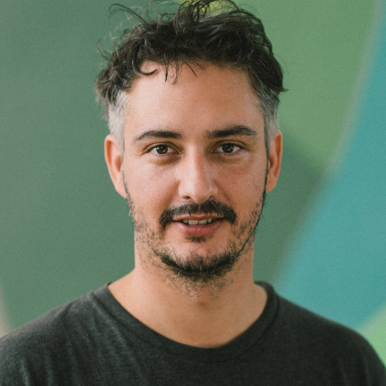 Ein Mann mit kurzen dunklen Haaren und heller Gesichtsbehaarung steht vor einem unscharfen grün-blauen Hintergrund, trägt ein dunkles T-Shirt und blickt in die Kamera.