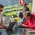 3 Männer an ihren Laptops. Im Hintergrund Aussicht aufs Berner Oberland und eine Schweizerfahne