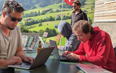 3 Männer an ihren Laptops. Im Hintergrund Aussicht aufs Berner Oberland und eine Schweizerfahne