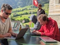 3 Männer an ihren Laptops. Im Hintergrund Aussicht aufs Berner Oberland und eine Schweizerfahne