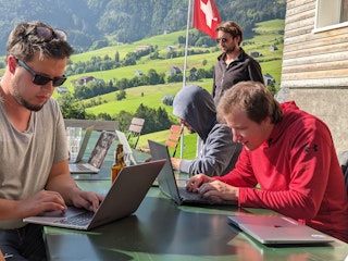 3 Männer an ihren Laptops. Im Hintergrund Aussicht aufs Berner Oberland und eine Schweizerfahne