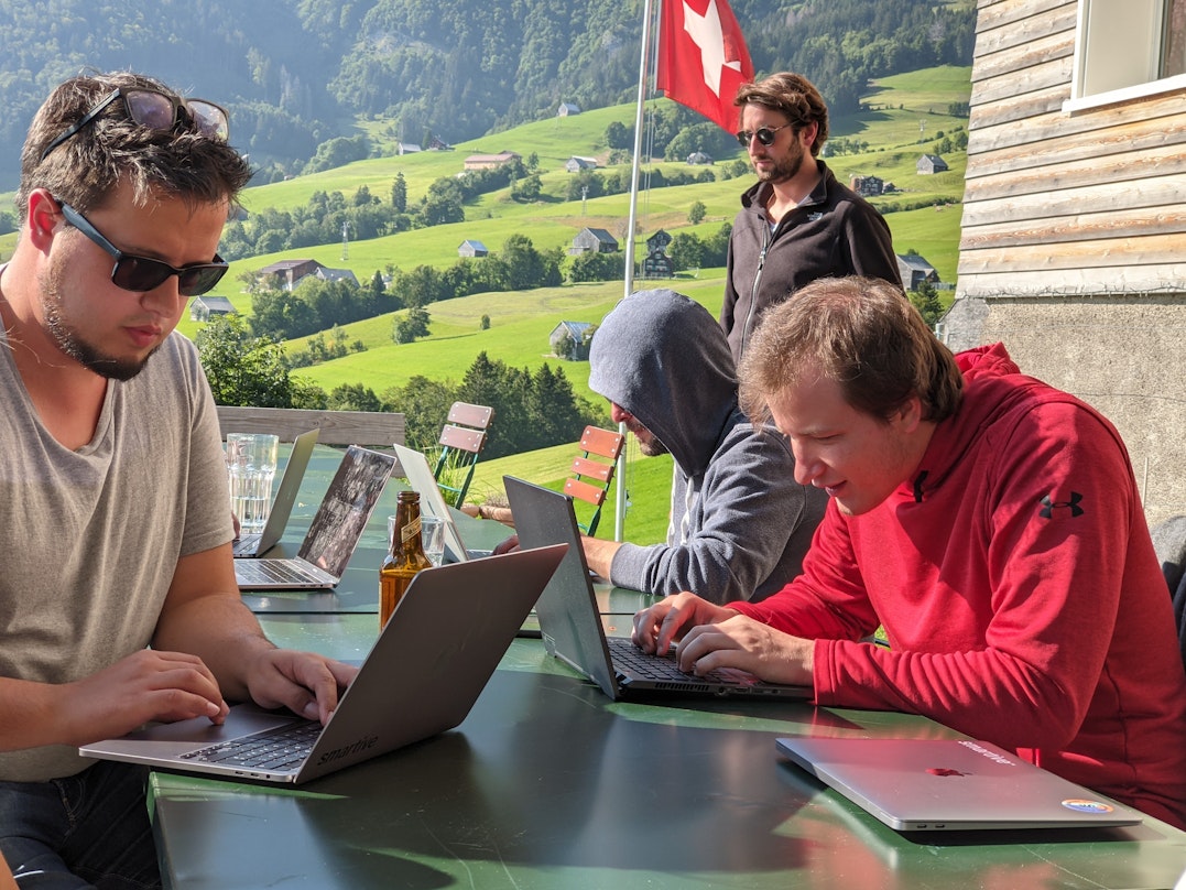 3 Männer an ihren Laptops. Im Hintergrund Aussicht aufs Berner Oberland und eine Schweizerfahne