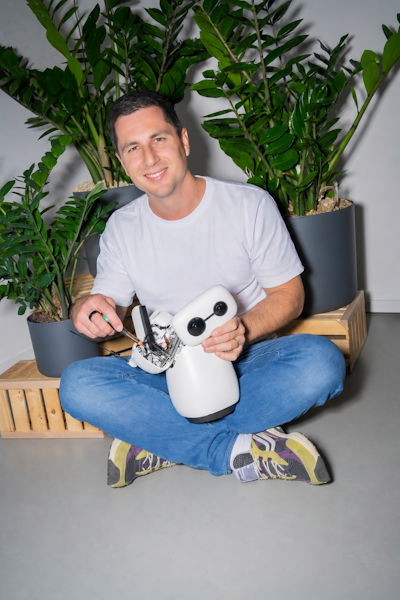 Ein Mann sitzt mit gekreuzten Beinen auf dem Boden, hält einen kleinen weissen Roboter in der Hand und repariert ihn, hinter ihm stehen zwei grosse Topfpflanzen.