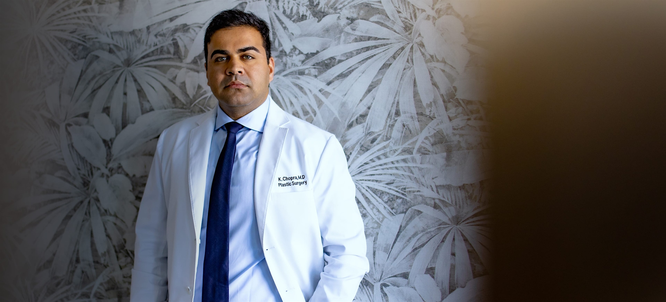 Dr. Chopra in a white coat, in front of a patterned wall