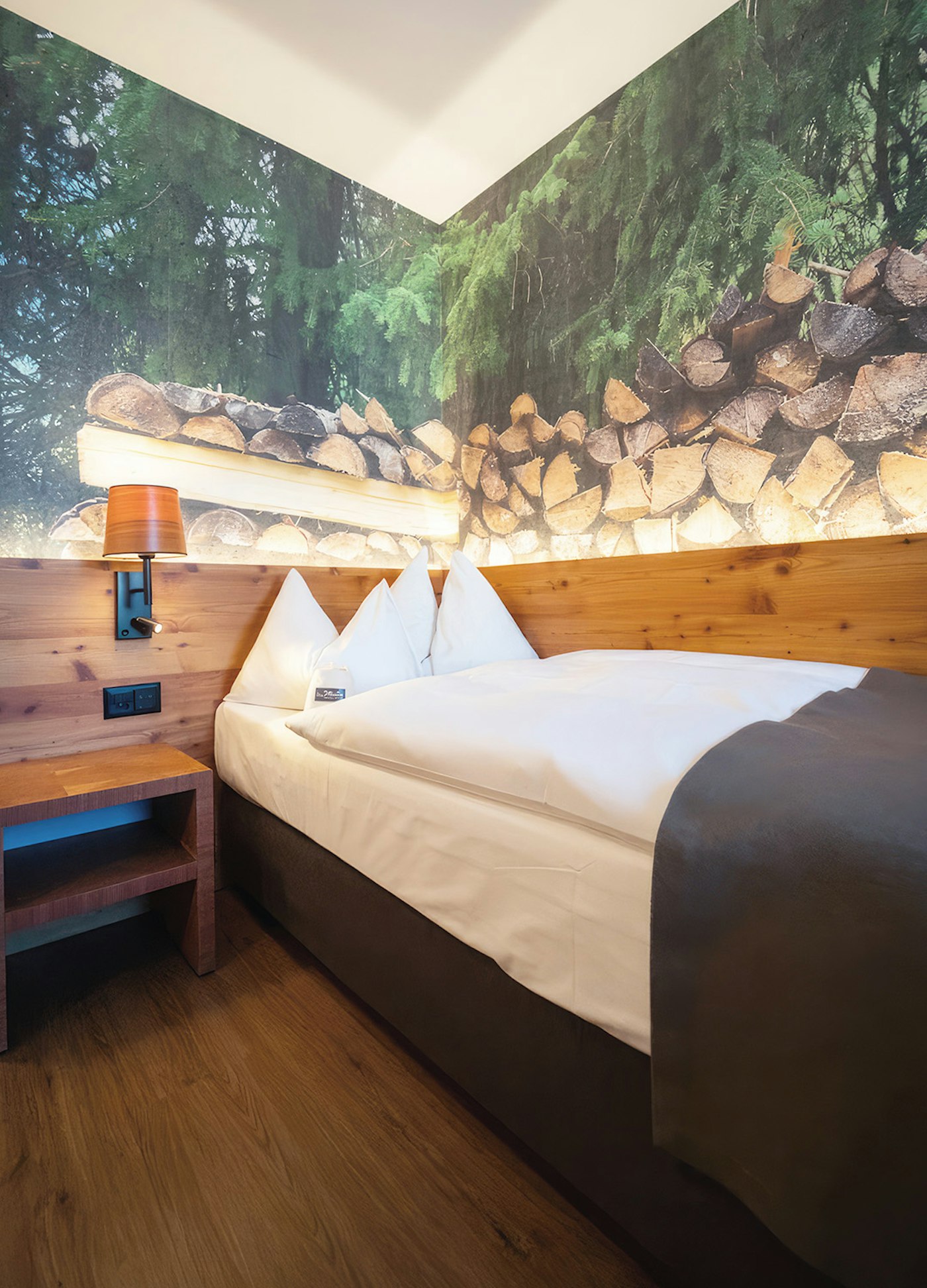 Einzelbett in der Ecke mit brauner Tagesdecke, Foto dahinter von einem Stapel Holz im Wald