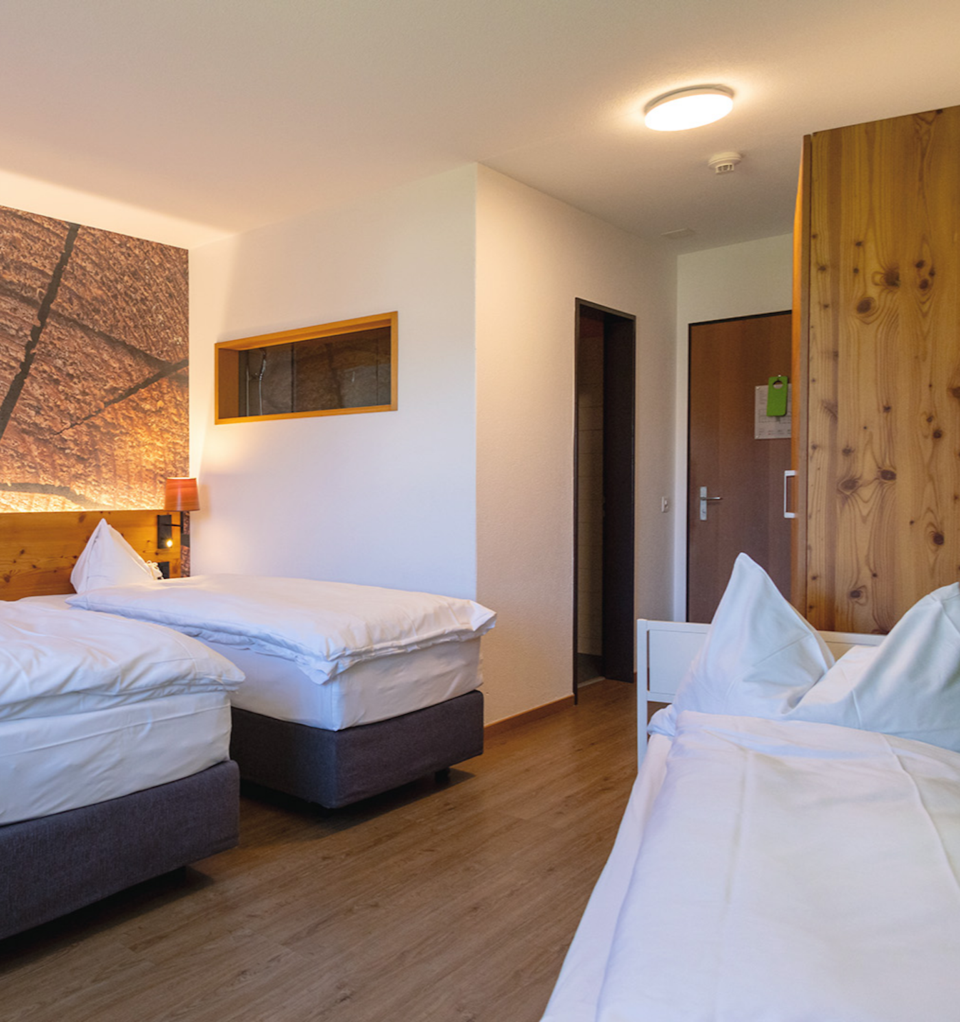 Zwei Betten stehen links mit Abstand an Wand, darüber Foto mit Holzstruktur, Beistellbett steht rechts neben dem Schrank, Sicht vom Fenster zur Türe und zum Badezimmer