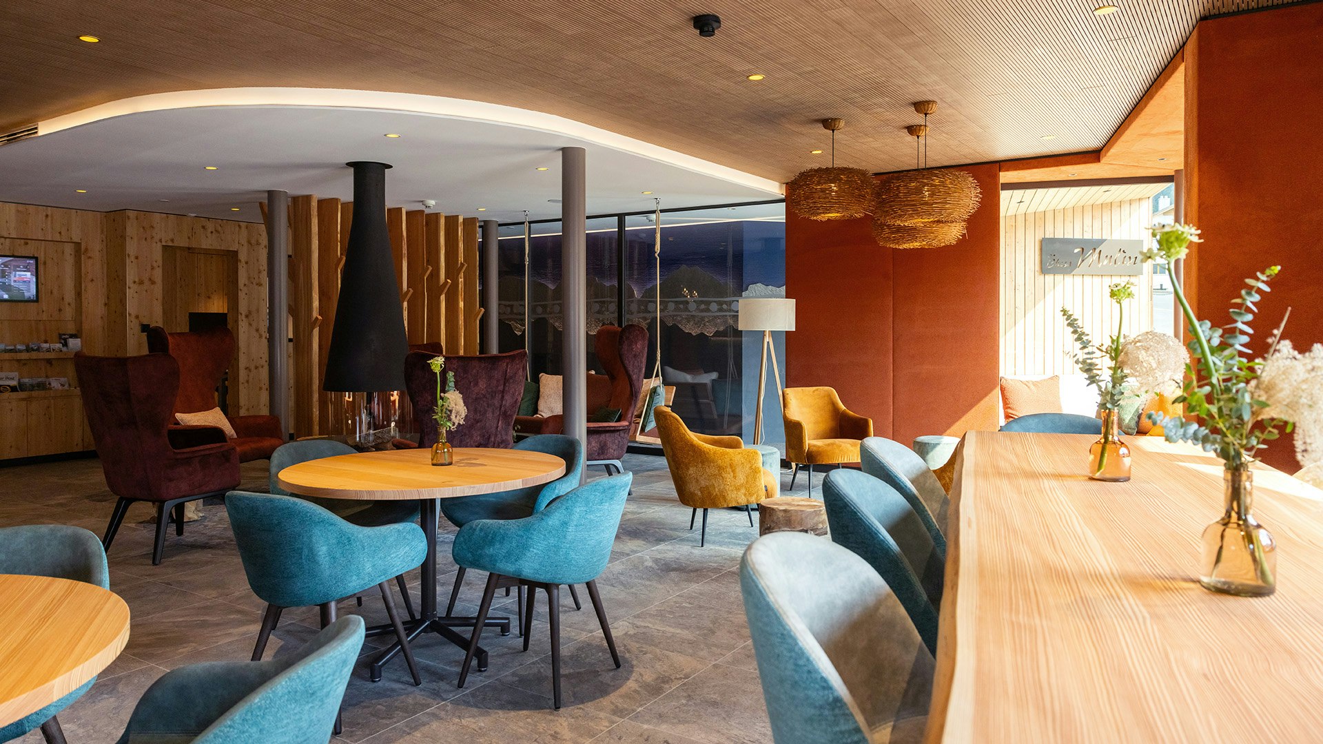 Eingangsbereich Lounge Blick auf Kamin und Lounge, im Vordergrund sind Holztische ische und Stühle in Türkis zu sehen