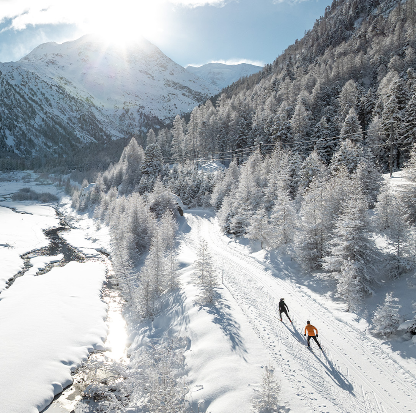 Zwei Langläufer in verschneiter Berglandschaft, bei Sonnenschein, links zugeschneiter Fluss