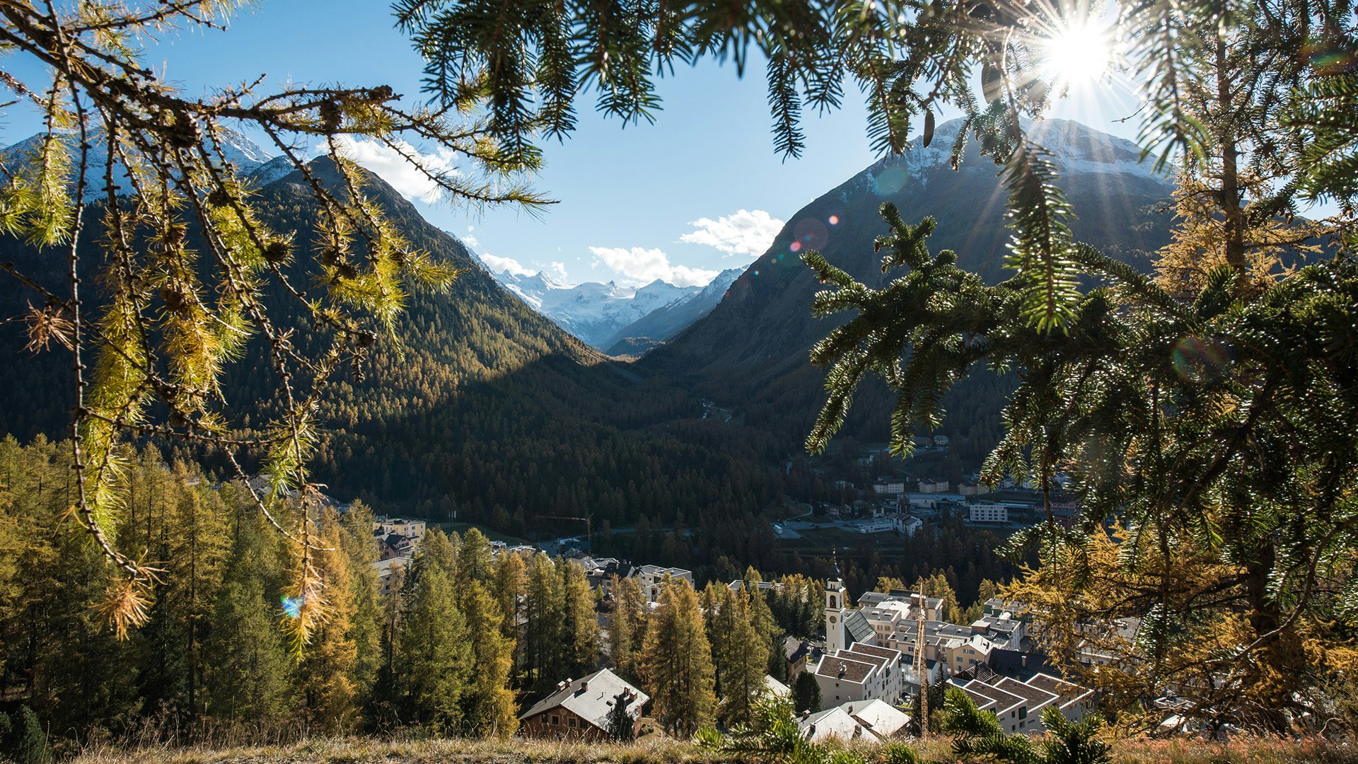 Das Dorf von oben von einem Hang aus gesehen, Sicht ins Rosegtal Berge und Sonnenstrahlen im Hintergrund