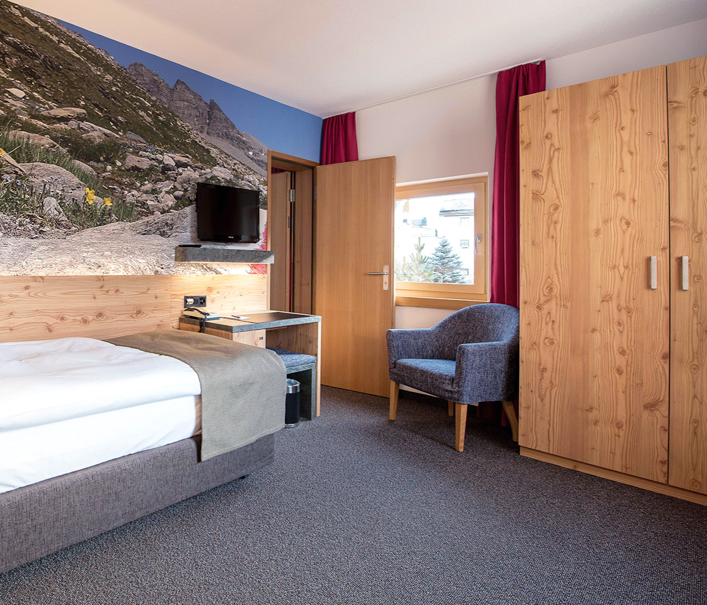 Links Einzelbett mit TV und Durchgang, daneben Sessel mit Holzschrank