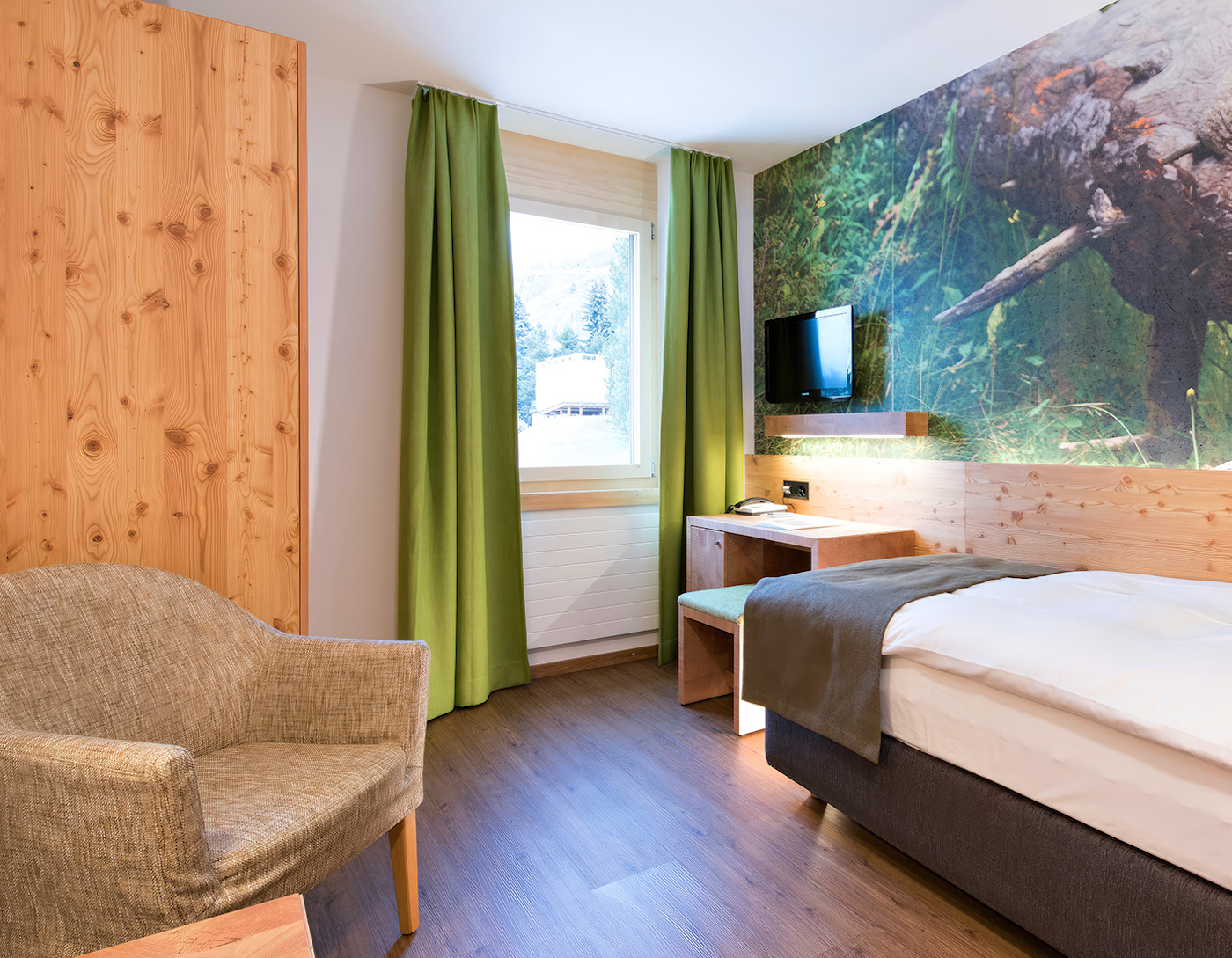 Blick auf Fenster mit grünen Vorhängen, rechts Holzschrank und Sessel, rechts Bettende mit Schreibtisch und TV