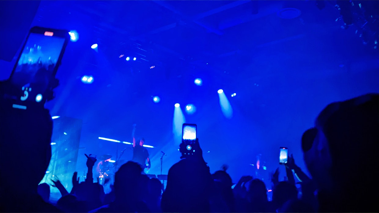 A performer on stage, bathed in blue light as crowd members record on their cellphones
