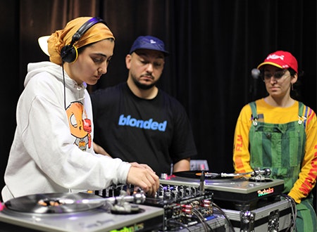 Female DJ performing while two others are observing.