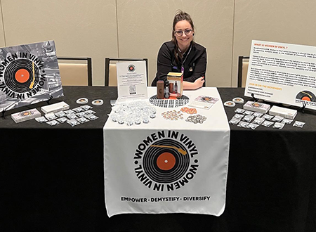 WIV-Event-2.png Jenn D'Eugenio sitting at a Women in Vinyl tradeshow booth.