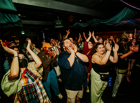 gRwX0Xcc-JUAN_UpHere_Day3_Night_137.png Crowd clapping and cheering for a band in a bar.