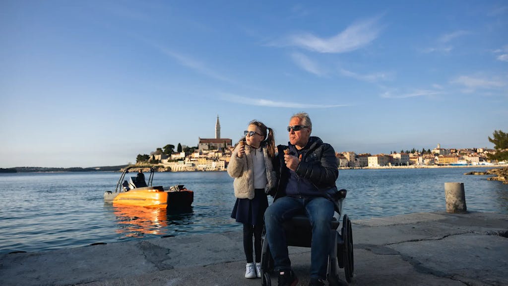 Grandfather in a wheelchair and granddaughter on the pier, with the Korkyra 650 Pro catamaran behind them.