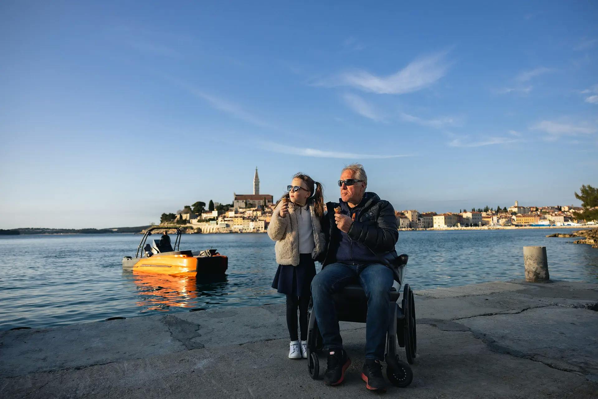 Grandfather in a wheelchair and granddaughter on the pier, with the Korkyra 650 Pro catamaran behind them.