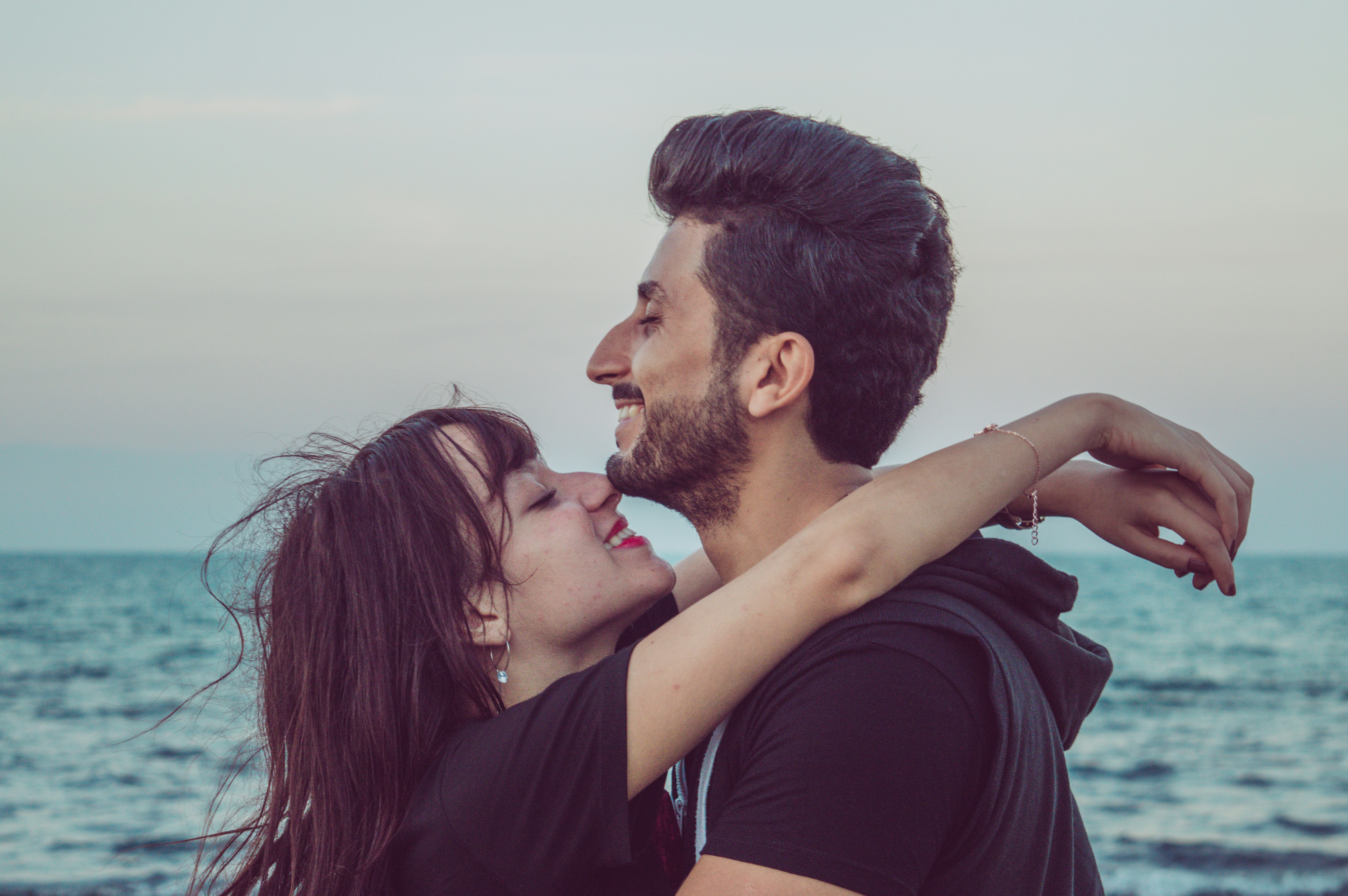 Smiling A young couple embraces and smiles at the beach. The woman, with windblown hair, looks up at the man while hugging him, and he smiles broadly, looking into the distance. The ocean and a clear sky form the serene background.