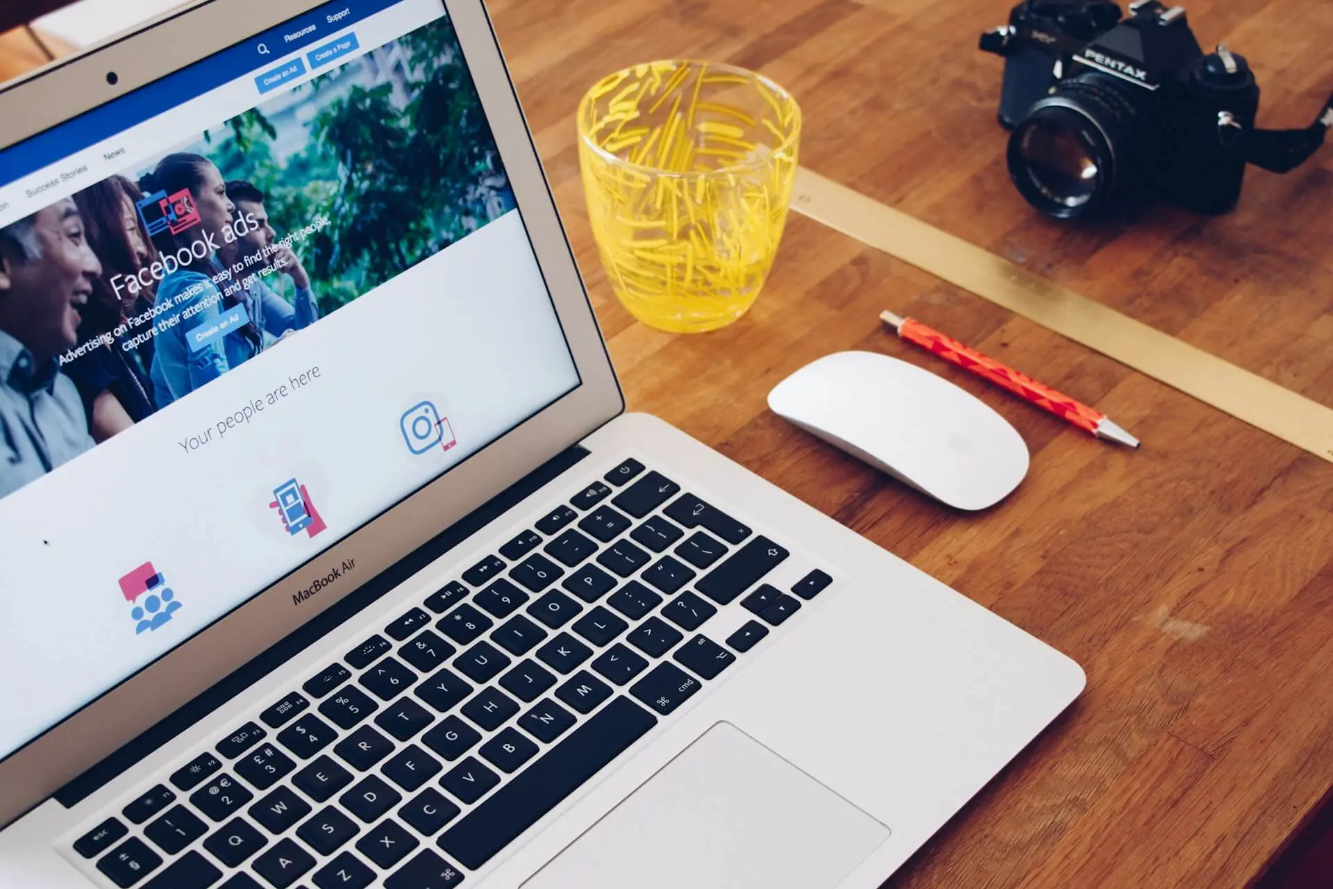 A MacBook Air displaying the Facebook Ads interface on a wooden desk, accompanied by a yellow glass, a red pencil, a wireless mouse, and a Pentax camera in the background.
