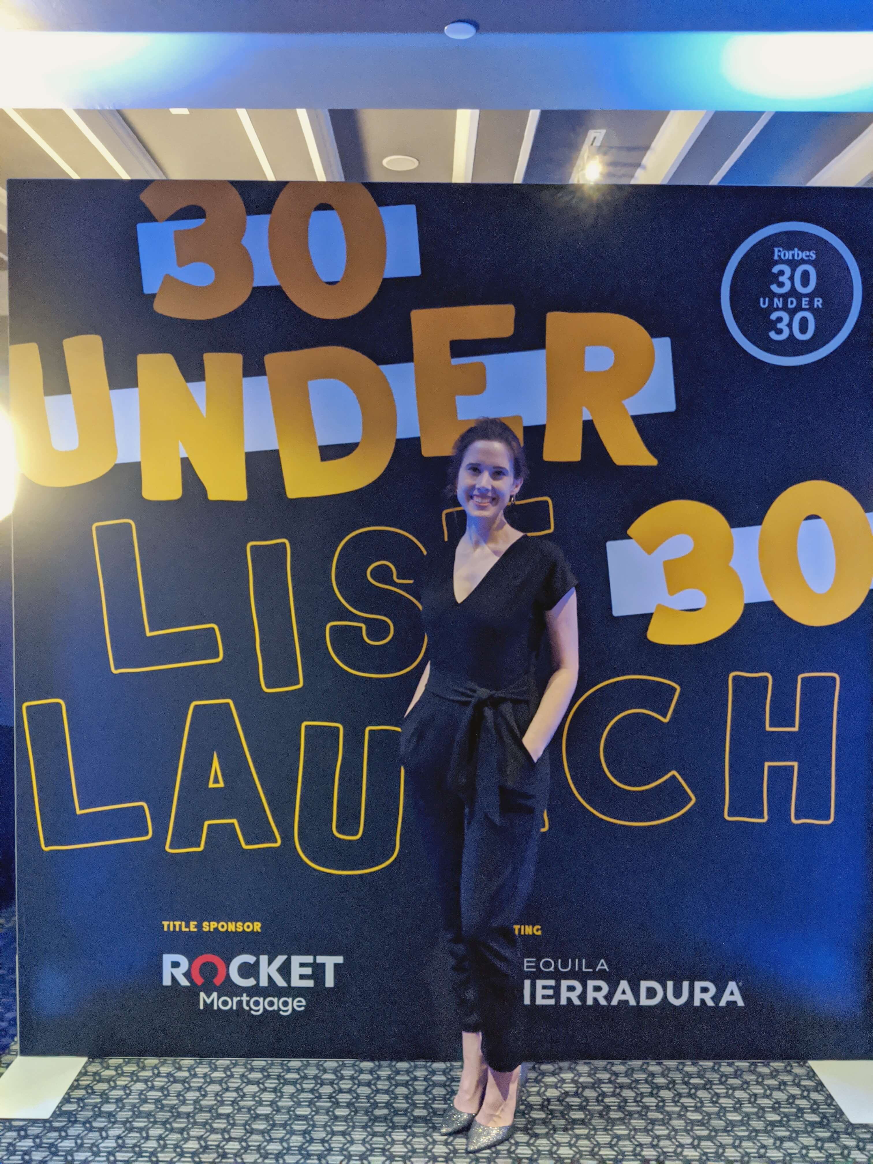 Smiling woman posing in front of a 'Forbes 30 Under 30 List Launch' banner at an event, representing young innovators and entrepreneurs celebrated for their achievements.