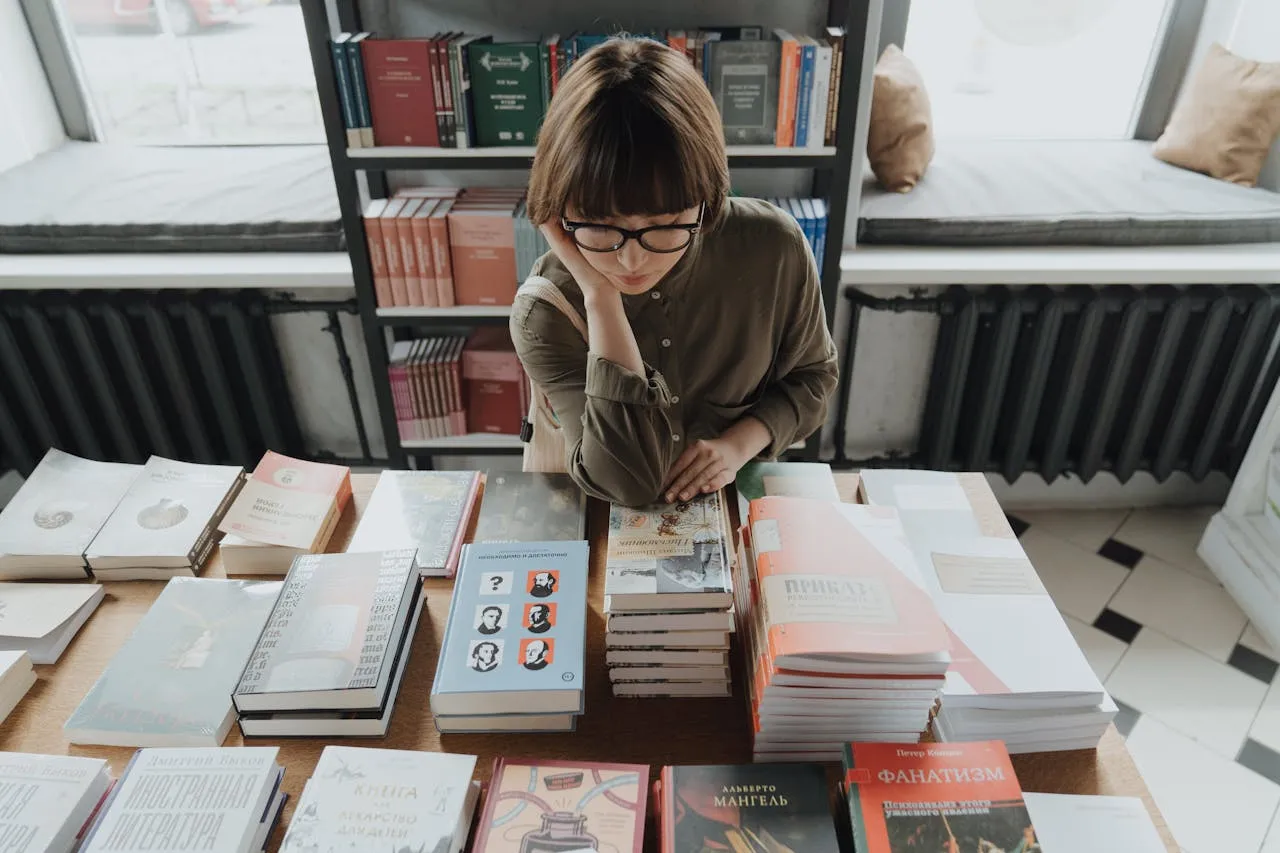 A young woman wearing glasses, leaning on her hand, browsing through books on a wooden table in a cozy bookstore with shelves of books in the background.