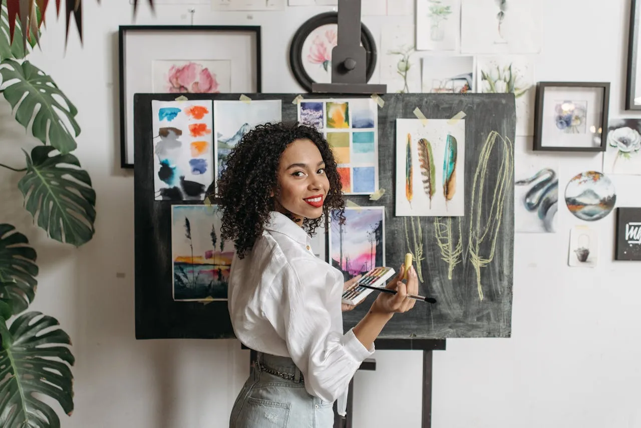 A smiling female artist with curly hair holding a paintbrush and palette, standing in front of a chalkboard easel displaying colorful art samples and sketches in a creative studio.
