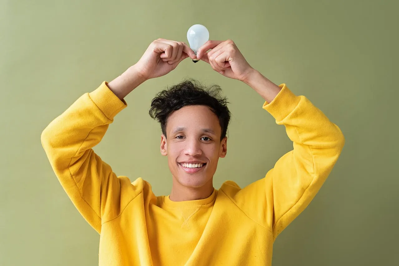 Smiling young man in a yellow sweater holding a lightbulb above his head against a green background, symbolizing creativity, innovation, and bright ideas in a vibrant and playful context.