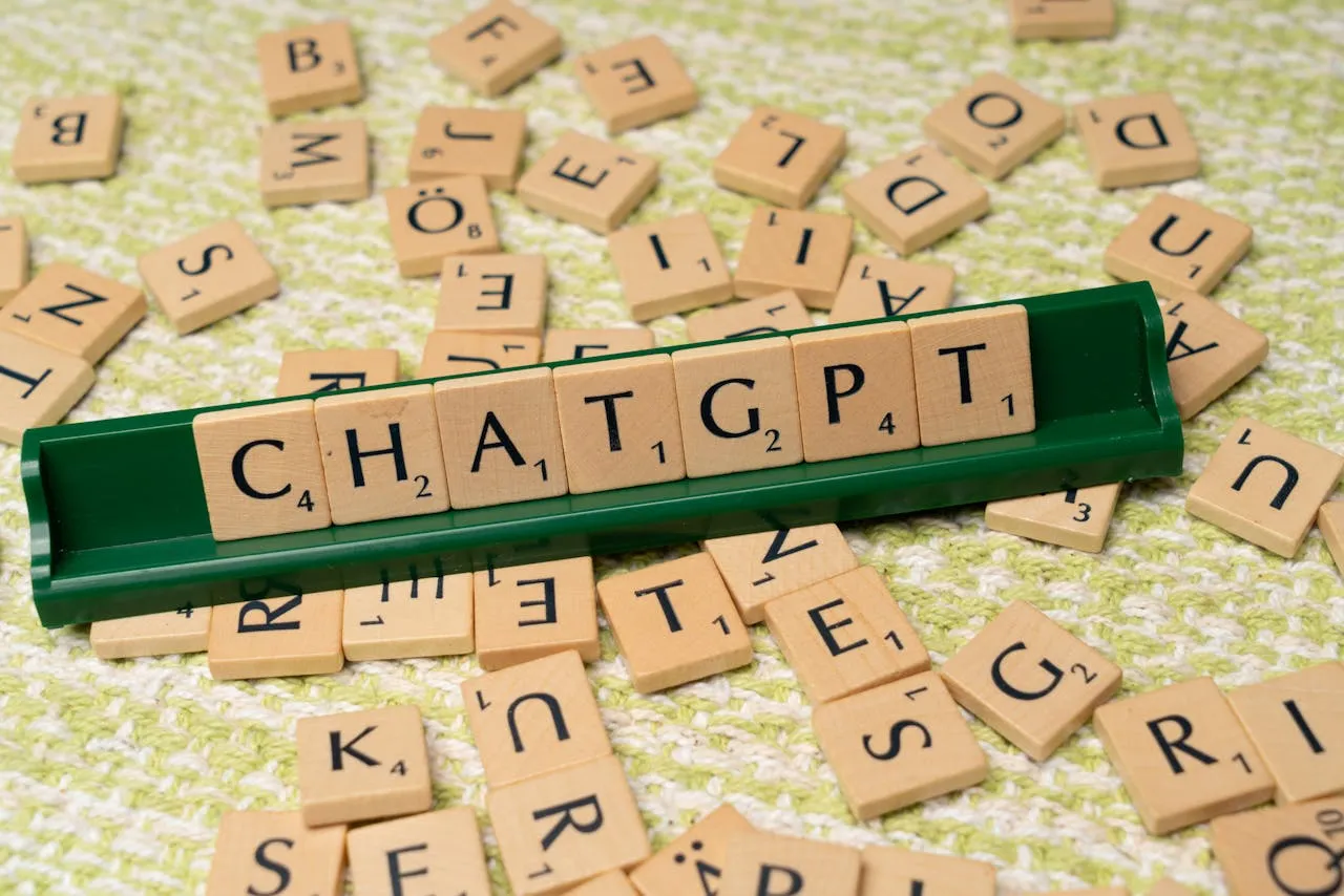 Scrabble-style letter tiles spelling out "CHATGPT" on a green tile holder, surrounded by scattered wooden tiles on a textured surface, representing AI, language models, and word games.