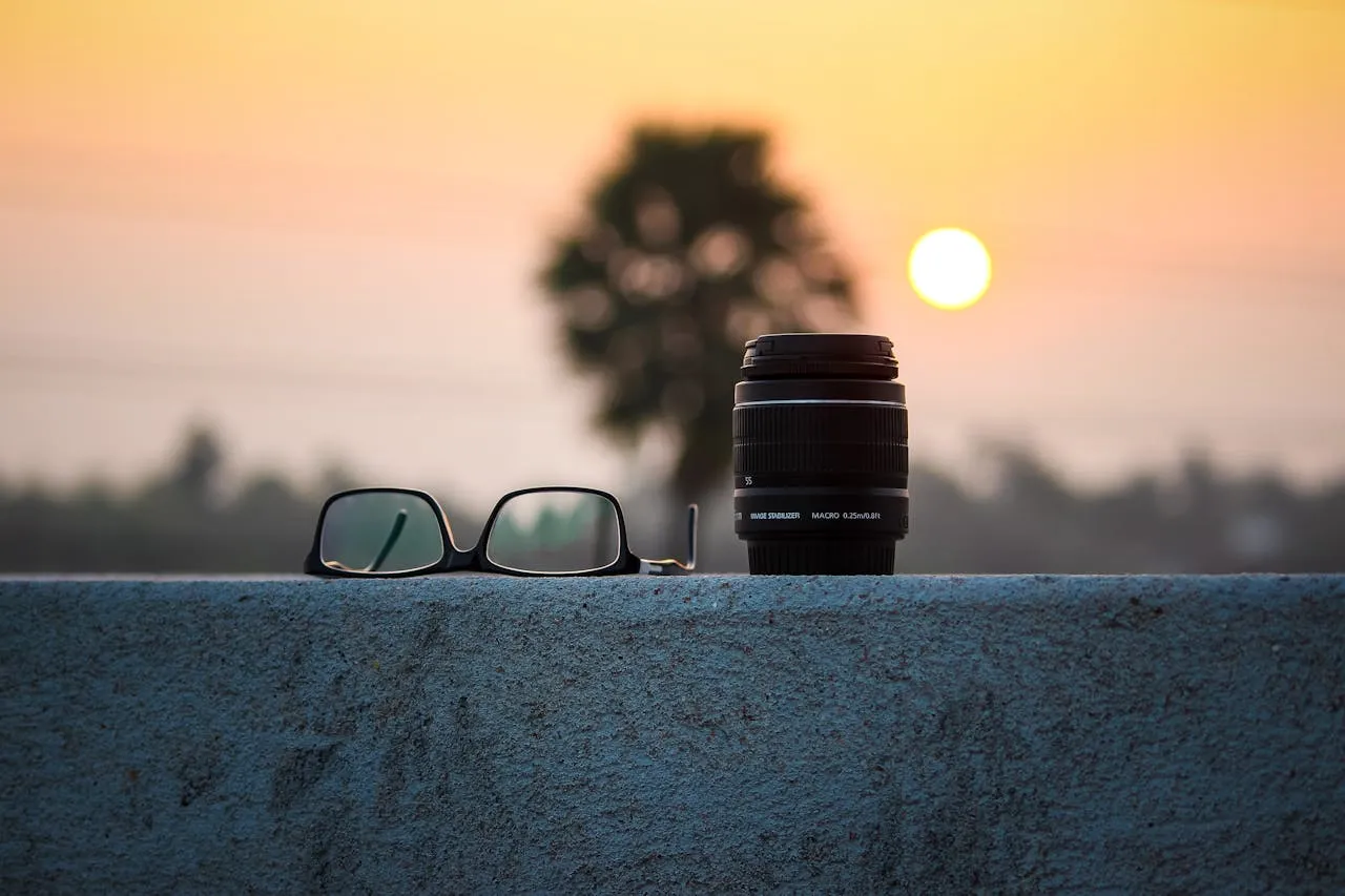 Camera lens and eyeglasses on a concrete ledge during sunset with a blurred tree and golden sun in the background.